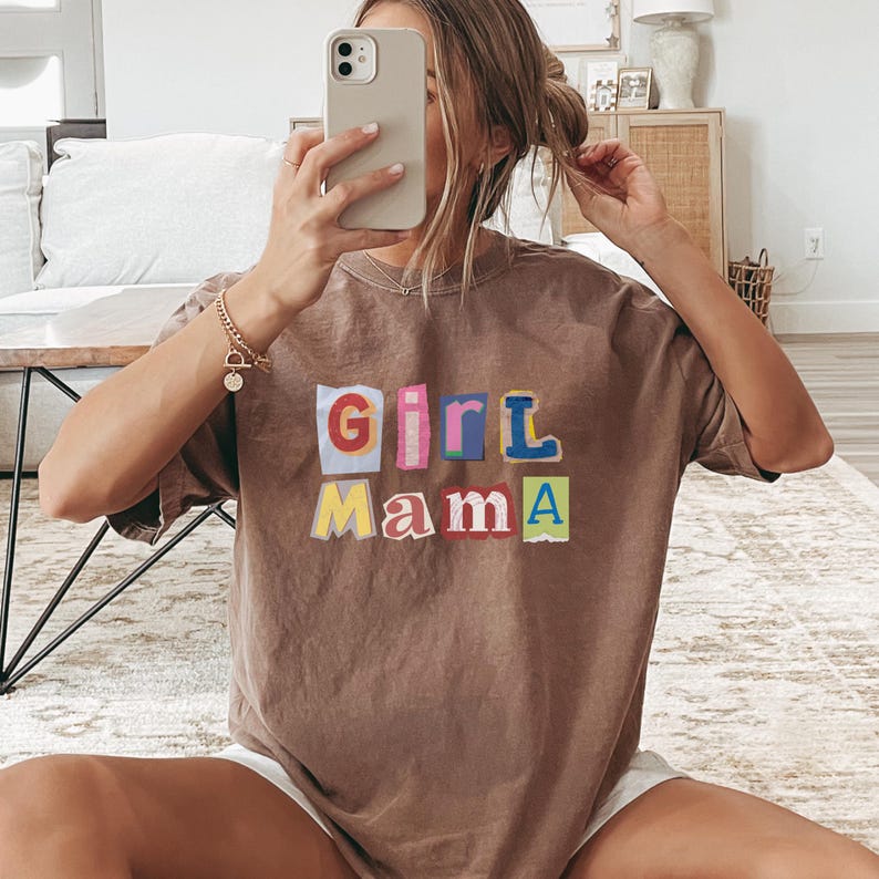 May include: Brown t-shirt with the text "Girl Mama" in colorful, cut-out style letters. The shirt is a relaxed fit and appears to be made of soft material. The overall aesthetic is casual and comfortable.