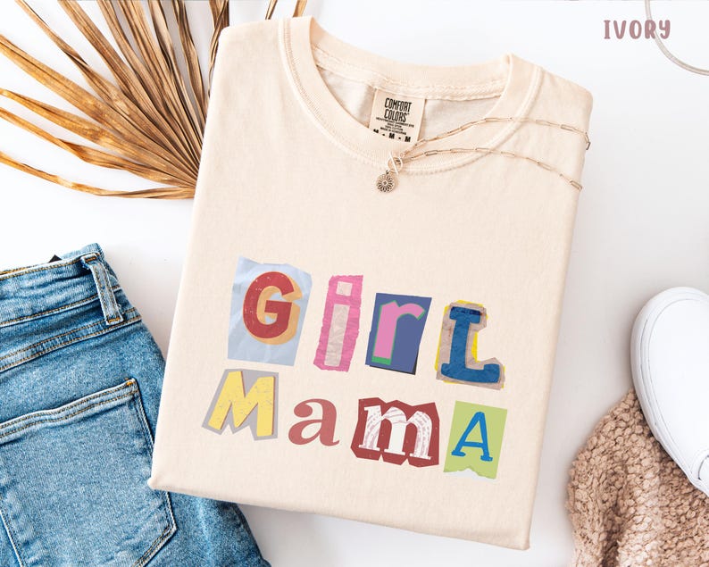 May include: Ivory-colored t-shirt with the words "Girl Mama" in a colorful, cut-out style. The shirt is folded, with a necklace and a pair of blue jeans visible in the background. The shirt is made of a soft material.