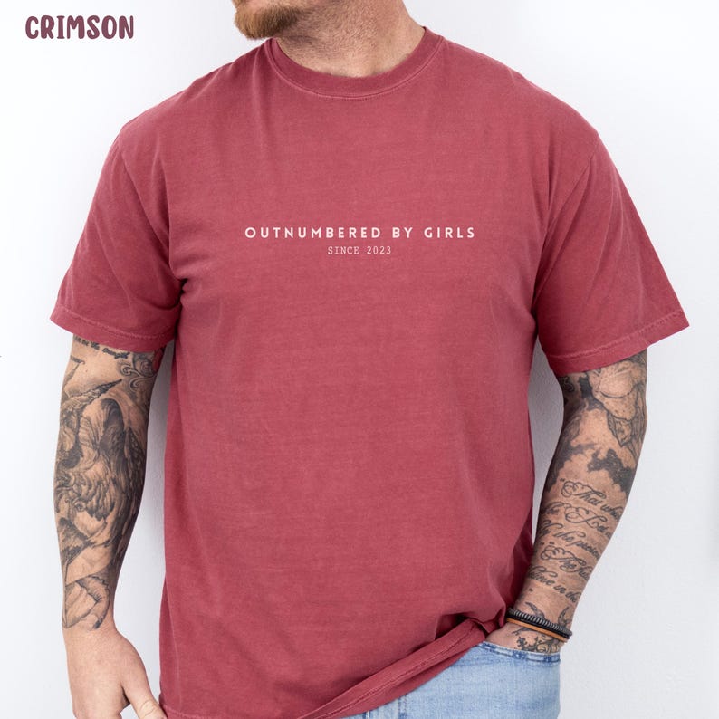 Może przedstawiać: Karmazynowy t-shirt z kr&oacute;tkim rękawem z białym napisem "OUTNUMBERED BY GIRLS" wyśrodkowanym na klatce piersiowej, z mniejszym tekstem "SINCE 2023" umieszczonym bezpośrednio pod gł&oacute;wnym hasłem, zapisanym wyraźną czcionką bezszeryfową.