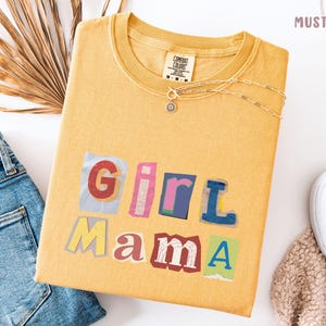 May include: Mustard-colored t-shirt with the words "Girl Mama" in a colorful, cut-out style. The shirt is paired with blue jeans, a silver necklace, and white sneakers. The shirt has a tag that says "Comfort Colors".