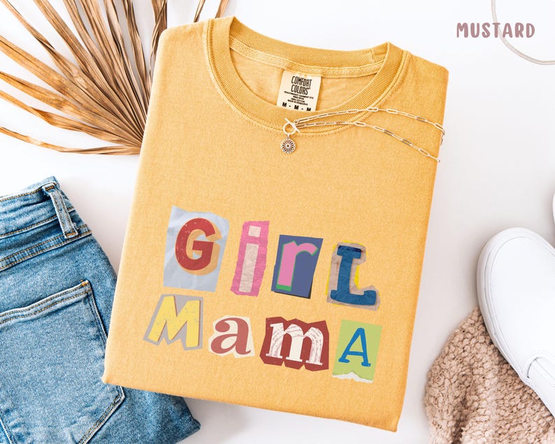 May include: Mustard-colored t-shirt with the words "Girl Mama" in a colorful, cut-out style. The shirt is paired with blue jeans, a silver necklace, and white sneakers. The shirt has a tag that says "Comfort Colors".