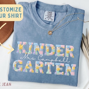 Puede incluir: Una camiseta color azul vaquero con el texto "KINDER GARTEN" en letras de bloque con un patrón de patchwork floral y de cuadros vichy. El nombre "Mrs. Campbell" está impreso en cursiva blanca entre las palabras. Una etiqueta dice "Comfort Colors".