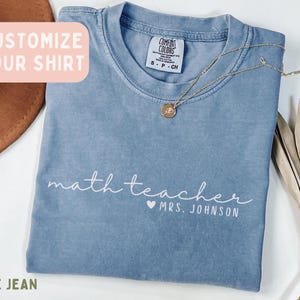 Puede incluir: Una camiseta color azul jean con cuello redondo y etiqueta de la marca Comfort Colors. La prenda presenta texto en blanco que dice math teacher con un símbolo de corazón y el nombre MRS. JOHNSON impreso debajo en una fuente clara.