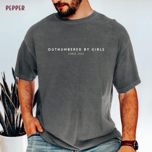 Może przedstawiać: Ciemnoszary t-shirt z kr&oacute;tkim rękawem z białym napisem "OUTNUMBERED BY GIRLS" wyśrodkowanym na klatce piersiowej oraz mniejszym tekstem "SINCE 2023" bezpośrednio pod nim. Koszulka ma okrągły dekolt i luźny, swobodny kr&oacute;j.