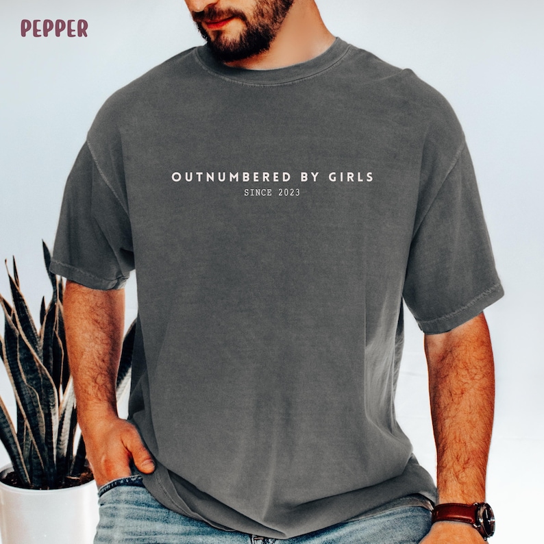 Może przedstawiać: Ciemnoszary t-shirt z kr&oacute;tkim rękawem z białym napisem "OUTNUMBERED BY GIRLS" wyśrodkowanym na klatce piersiowej oraz mniejszym tekstem "SINCE 2023" bezpośrednio pod nim. Koszulka ma okrągły dekolt i luźny, swobodny kr&oacute;j.