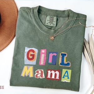 May include: Moss green t-shirt with the words "Girl Mama" in a colorful, cut-out style. A gold necklace with a pendant rests on the shirt. A brown hat and dried plant are in the background.