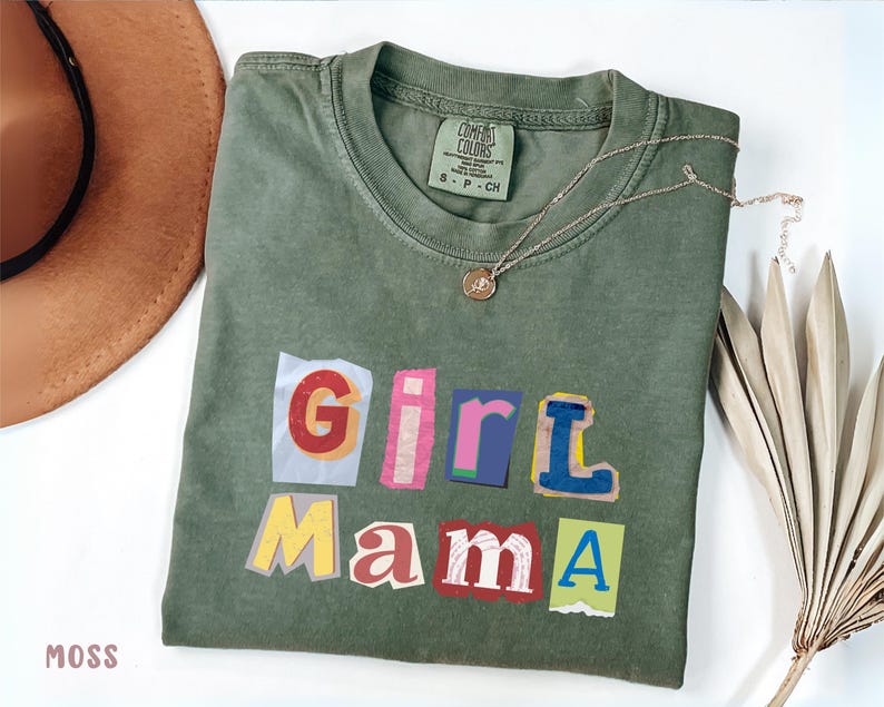May include: Moss green t-shirt with the words "Girl Mama" in a colorful, cut-out style. A gold necklace with a pendant rests on the shirt. A brown hat and dried plant are in the background.