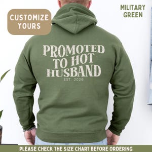 May include: A military green hooded sweatshirt featuring the text PROMOTED TO HOT HUSBAND EST. 2026 printed in a distressed white font on the back. The hoodie includes a hood and long sleeves, with the text CUSTOMIZE YOURS and MILITARY GREEN visible.
