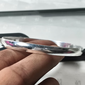 May include: A silver bangle bracelet with a smooth, rounded design. The bracelet is a solid band of polished metal, reflecting light. The jewelry is held in a hand, showcasing its simple, elegant style.