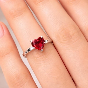 Puede incluir: Un anillo de plata con una piedra preciosa roja en forma de corazón. El anillo está engastado con cuatro garras y se muestra en un dedo. El fondo es de un color suave y neutro.