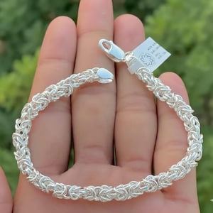 Peut inclure: Un bracelet argenté avec un motif tressé. Le bracelet est composé de petits maillons interconnectés, créant un aspect texturé. Il est doté d'un fermoir sécurisé pour un port facile. Le bracelet est présenté sur un fond vert flou.
