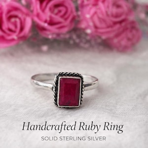 Könnte beinhalten: Ein handgefertigter Ring aus Sterlingsilber mit einem rechteckigen Rubin. Der Rubin hat eine tiefrote Farbe und ist in einem gedrehten, seilartigen Rahmen gefasst. Der Ring wird auf einer weißen Oberfläche mit rosa Blumen im Hintergrund präsentiert. Der Text lautet "Handcrafted Ruby Ring" und "Solid Sterling Silver."