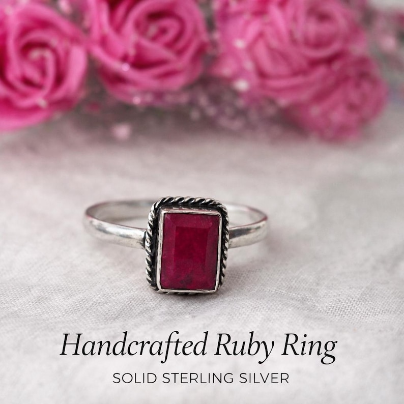 Puede incluir: Un anillo artesanal de plata de ley con una gema de rub&iacute; rectangular. El rub&iacute; es de color rojo intenso, engastado en un borde trenzado. El anillo se muestra sobre una superficie blanca con flores rosas en el fondo. El texto dice "Handcrafted Ruby Ring" y "Solid Sterling Silver."