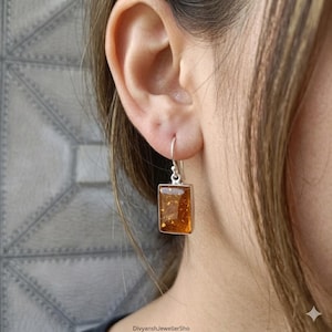 May include: A silver rectangular earring with a warm amber-colored stone. The amber has flecks of gold throughout. The earring is hanging from a silver hook. The earring is shown on a person's ear.