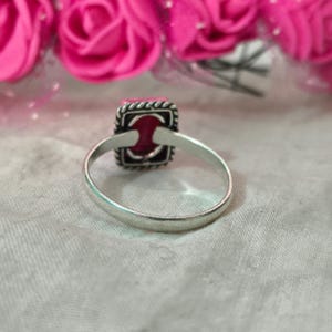 Puede incluir: Un anillo de plata con una piedra preciosa cuadrada de color rojo oscuro. La piedra preciosa est&aacute; engastada en un marco cuadrado con un borde decorativo. El anillo est&aacute; sobre una superficie gris claro, con flores rosas en el fondo.