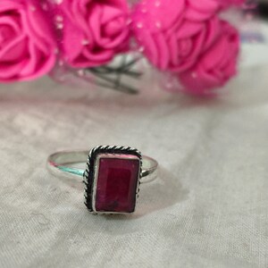 Puede incluir: Anillo de plata con una gema rectangular de color rojo intenso. La gema est&aacute; engastada en un marco met&aacute;lico retorcido y decorativo. Flores rosas en el fondo.