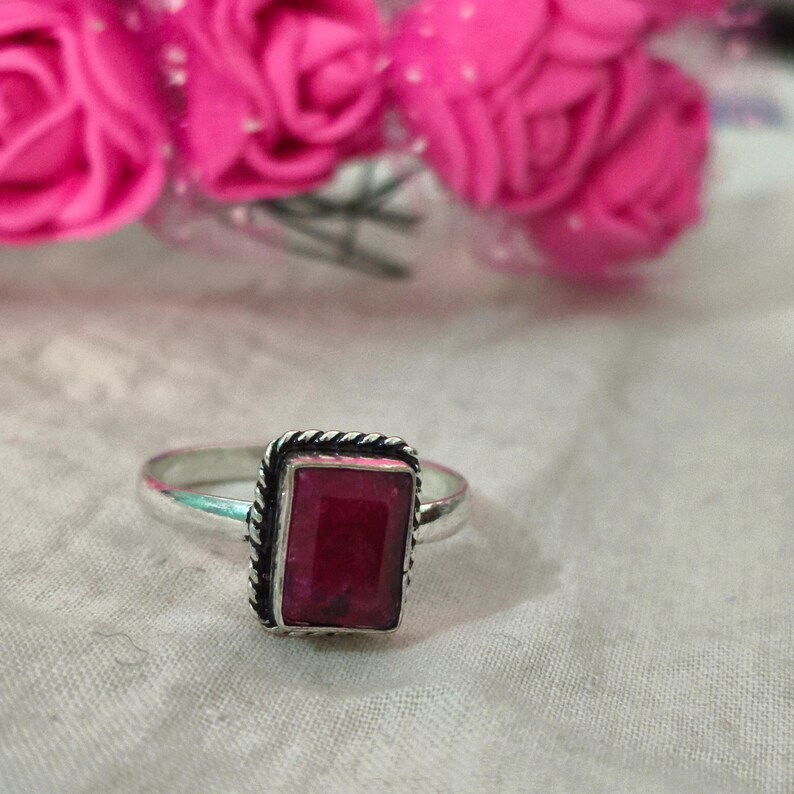 Puede incluir: Anillo de plata con una gema rectangular de color rojo intenso. La gema est&aacute; engastada en un marco met&aacute;lico retorcido y decorativo. Flores rosas en el fondo.