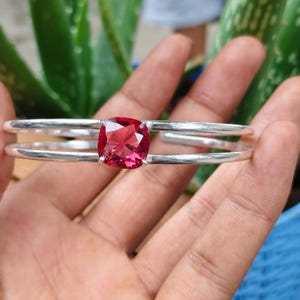 Può includere: Un bracciale rigido in argento con una pietra preziosa centrale di colore rosa intenso a taglio cuscino. Il bracciale è composto da due fasce d'argento lisce e parallele che si incontrano sull'incastonatura della pietra sfaccettata rosa.
