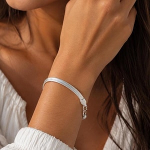Puede incluir: Una pulsera de cadena de espiga en tono plateado con un diseño de eslabones planos y flexibles con un acabado metálico pulido. La pulsera incluye un pequeño y delicado cierre de mosquetón y una cadena extensora corta con un aro.