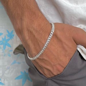 Könnte beinhalten: Ein silbernes Panzerkettenarmband. Das Armband besteht aus ineinandergreifenden Gliedern und wird am Handgelenk getragen. Das Bild zeigt das Armband vor einem neutralen Hintergrund, der seinen metallischen Glanz und sein Design hervorhebt.
