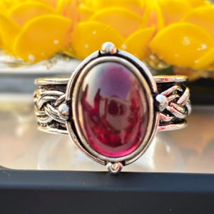 May include: A silver ring featuring a large, oval-shaped, deep red cabochon gemstone set in the centre. The band of the ring displays a detailed, braided metal texture that wraps around the finger, providing a contrast to the smooth stone.