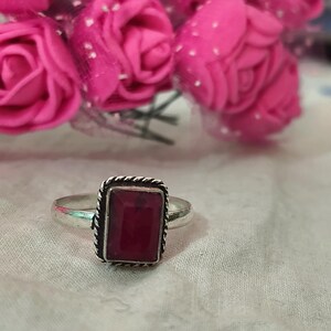Puede incluir: Un anillo de plata con una piedra preciosa rectangular de color rojo intenso. La piedra preciosa est&aacute; engastada en un marco cuadrado con un dise&ntilde;o de cuerda trenzada. Rosas rosas en el fondo.