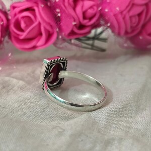 Puede incluir: Un anillo de plata con una piedra preciosa rosa oscuro rectangular. La piedra est&aacute; engastada en un marco cuadrado con un borde decorativo. El anillo se exhibe sobre una tela blanca, con flores rosas en el fondo.