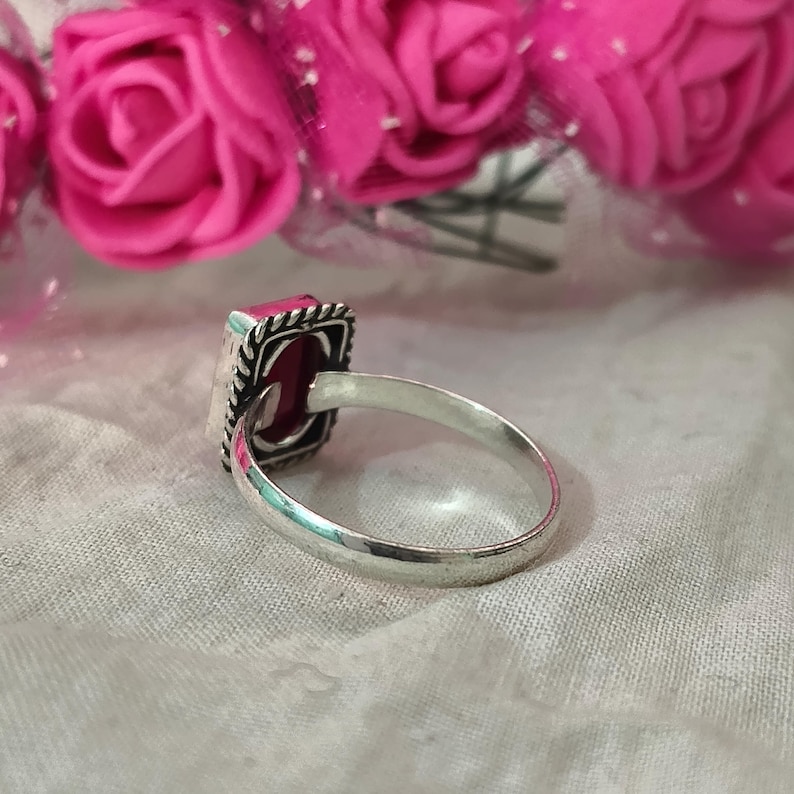Puede incluir: Un anillo de plata con una piedra preciosa rosa oscuro rectangular. La piedra est&aacute; engastada en un marco cuadrado con un borde decorativo. El anillo se exhibe sobre una tela blanca, con flores rosas en el fondo.