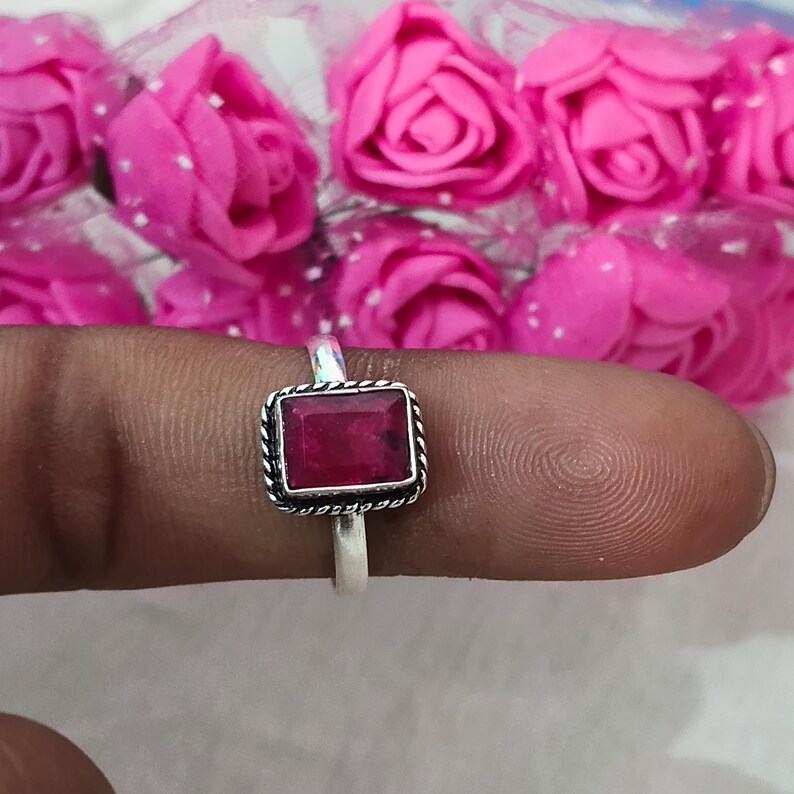 Puede incluir: Un anillo de plata con una piedra preciosa rectangular de color rojo intenso. La piedra preciosa est&aacute; engastada en un borde decorativo en forma de cuerda. El anillo se muestra sobre un fondo de rosas rosas.