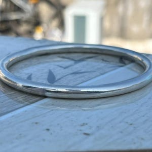 May include: A silver-colored, round bangle bracelet. The bracelet has a smooth, polished surface and a simple, minimalist design. The bangle is approximately 2.5 inches in diameter, suitable for everyday wear.