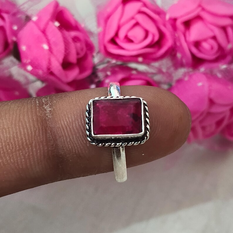 Puede incluir: Un anillo de plata con una piedra preciosa rectangular de color rojo intenso. La piedra preciosa est&aacute; engastada en un marco cuadrado con un dise&ntilde;o de cuerda trenzada. El anillo se muestra sobre un fondo de flores rosas.