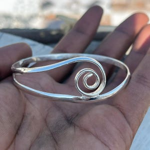 Könnte beinhalten: Ein silberfarbenes Armband mit einem Spiraldesign. Das Armband ist im Bangle-Stil gehalten und hat eine glatte, polierte Oberfläche. Die Spirale ist mittig auf dem Armband platziert und verleiht dem Schmuck eine einzigartige Note.