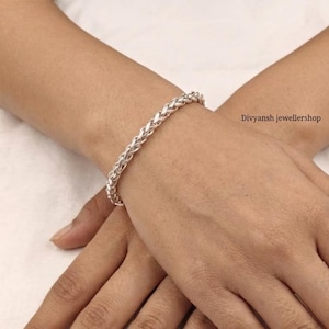 Pode incluir: Uma pulseira prateada com um design trançado. A pulseira está em um pulso, e a imagem é um close-up. A pulseira é feita de um metal brilhante e tem uma superfície texturizada. O texto "Divyansh jewellershop" é visível.