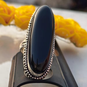 Puede incluir: Anillo de plata con una gran piedra ovalada negra pulida. El anillo tiene un borde detallado con cuentas. Flores amarillas en el fondo añaden un toque de color a la imagen.