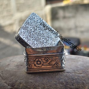 May include: A square silver box with an intricate floral and geometric pattern engraved on the lid and sides. The lid is partially open, revealing the metallic interior. The text Dwyanshjewellerysop is visible across the surface of the lid.