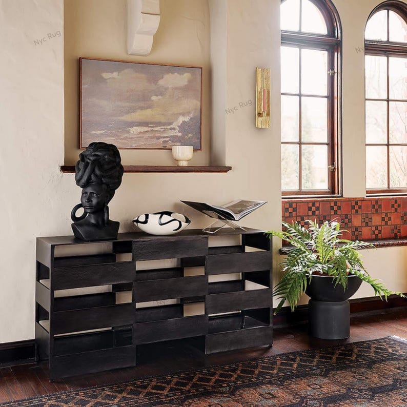 Pu&ograve; includere: Una consolle in legno nero con un design geometrico, sormontata da una scultura a busto nero, una ciotola decorativa e un portariviste in metallo. Un dipinto e un'applique sono visibili sullo sfondo.