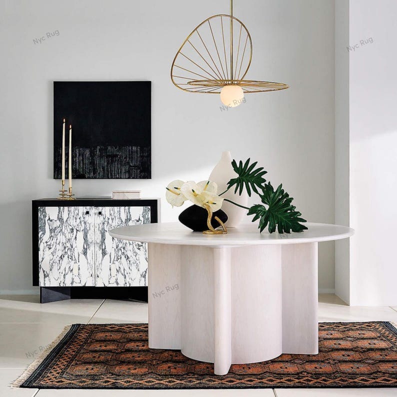 Pu&ograve; includere: Una sala da pranzo moderna con un tavolo rotondo bianco, una base a piedistallo unica, un mobile bianco e nero e un lampadario dorato. La stanza &egrave; decorata con un tappeto e vasi con piante.