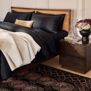 Pu&ograve; includere: Una scena di camera da letto con un letto con biancheria da letto nera, inclusi cuscini e un piumino. Una coperta color crema &egrave; drappeggiata sui piedi del letto. Un comodino in legno con piano in marmo contiene un vaso di fiori e un bicchiere d'acqua. Un tappeto fantasia &egrave; sul pavimento.