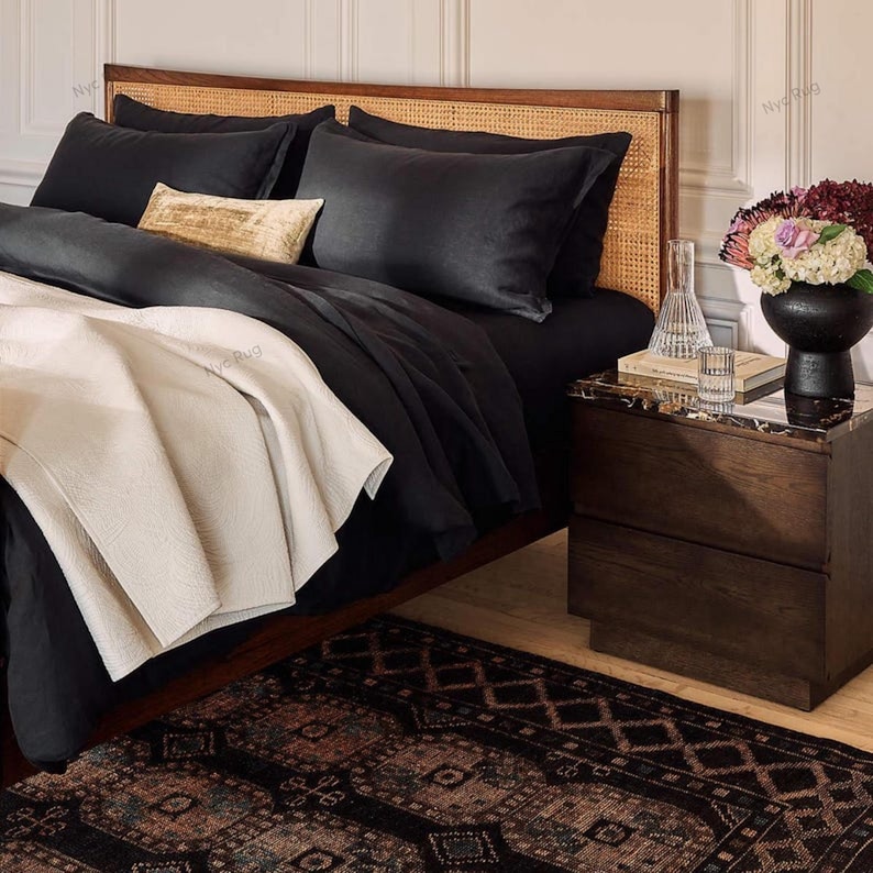 Pu&ograve; includere: Una scena di camera da letto con un letto con biancheria da letto nera, inclusi cuscini e un piumino. Una coperta color crema &egrave; drappeggiata sui piedi del letto. Un comodino in legno con piano in marmo contiene un vaso di fiori e un bicchiere d'acqua. Un tappeto fantasia &egrave; sul pavimento.