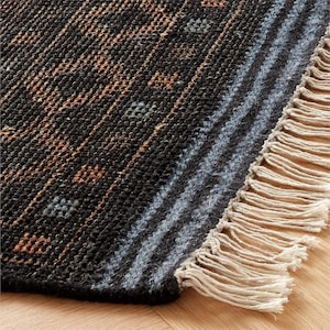 Pu&ograve; includere: Primo piano di un tappeto tessuto con un motivo geometrico. Il tappeto presenta uno sfondo scuro con strisce diagonali nelle tonalit&agrave; del marrone, blu e grigio. Il bordo del tappeto ha una frangia color beige chiaro.