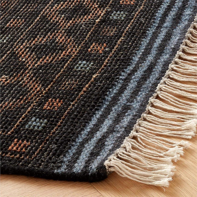 Pu&ograve; includere: Primo piano di un tappeto tessuto con un motivo geometrico. Il tappeto presenta uno sfondo scuro con strisce diagonali nelle tonalit&agrave; del marrone, blu e grigio. Il bordo del tappeto ha una frangia color beige chiaro.