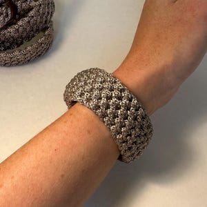 May include: A woven, taupe-colored bracelet is worn on a person's wrist. A matching belt with a brown buckle is coiled nearby. The bracelet and belt have a textured, open weave design. The bracelet is approximately 2 inches wide.