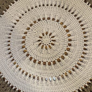 May include: A round, hand-crocheted rug in shades of beige and brown. The rug features concentric circles with a detailed, textured pattern. The outer border is a darker brown, while the inner sections are lighter, creating a visual contrast.