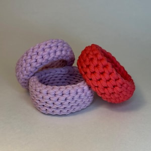 May include: Three handmade crochet baskets in shades of purple and red. The baskets are round with a textured, woven appearance. The colors range from light lavender to a vibrant red-orange.