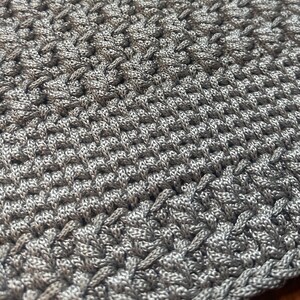 May include: Close-up of a hand-crocheted item in a light gray color. The texture is dense and intricate, with a woven appearance. The yarn appears to be a soft, possibly cotton or acrylic blend. The overall effect is of a handmade textile.