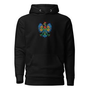 May include: Black hooded sweatshirt featuring a colorful embroidered Polish eagle emblem. The eagle design incorporates red, yellow, blue, and green floral patterns. The sweatshirt has a front pocket and a drawstring hood.