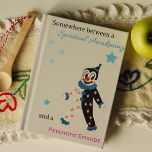 以下が含まれることがあります： 「Somewhere between a Spiritual Awakening and a Psychotic Episode」というテキストと漫画の道化師のイラストが描かれた本の表紙。 道化師は黒色で、ピンクのダイヤモンドのアクセントと青い襟が付いています。 薄い青色の星がテキストの周りに散らばっています。