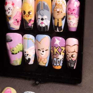 Op de afbeelding: Een set van tien kunstnagels met handgeschilderde stripfiguren en ontwerpen. De nagels tonen diverse illustraties, waaronder dieren, een groene rups en de tekst SKZ CODE in het geel op een paarse achtergrond.