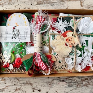 Può includere: Una scatola di cartone riempita con articoli per il fai da te a tema natalizio, tra cui una sagoma di Babbo Natale, fiocchi di neve in pizzo bianco, ritagli di tessuto rosso e verde, bottoni, perline, un cartoncino con spartito e una bobina.