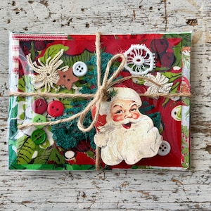 Può includere: Un kit artigianale rettangolare avvolto da spago, caratterizzato da una sagoma vintage di Babbo Natale, tessuto floreale rosso e verde, piccoli bottoni colorati, una stella in legno e dettagli in uncinetto bianco, sigillato in una busta trasparente.